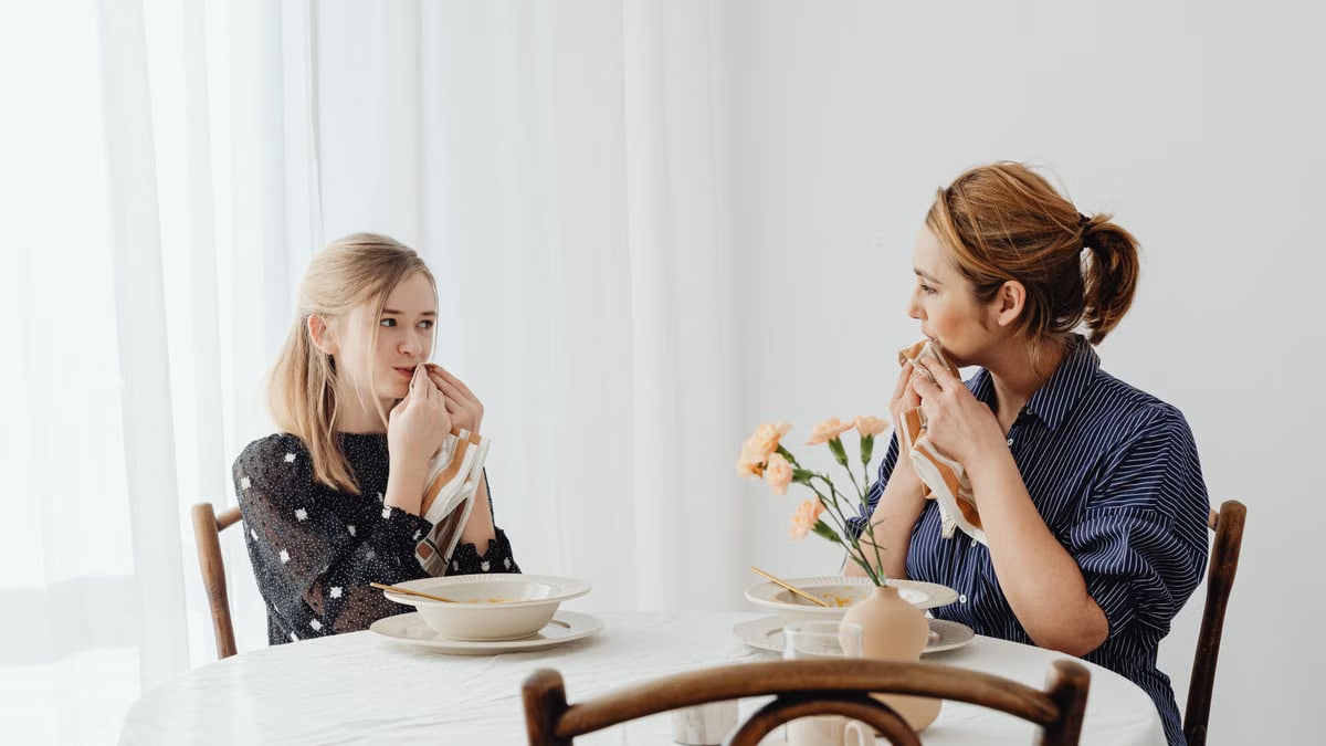 Mother and daughter share a cozy meal indoors, bonding and smiling at a dining table.