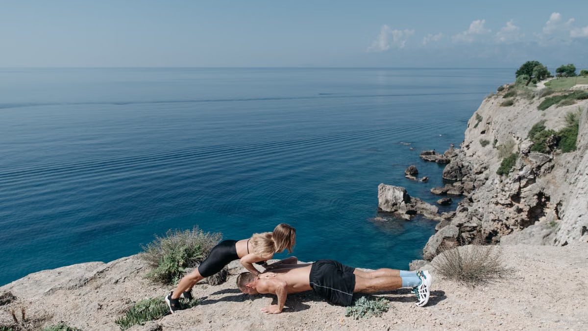 A couple performs a partner workout on a scenic cliffside, overlooking the ocean.